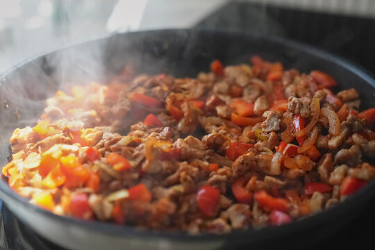 Meat pieces with red pepper and onion cooking in hot frying pan with steam rising