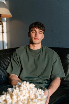 Young man sitting on sofa with bowl of popcorn watching movie in cozy living room for lifestyle background and streaming night banner use