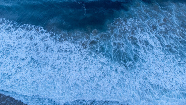 Aerial view of a powerful ocean wave breaking into intricate white foam, revealing rich blue water textures and dynamic natural movement.