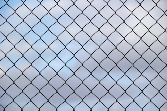 Chain-link fence silhouetted against a pale blue cloudy sky, creating a clean geometric pattern with atmospheric depth and a minimalist outdoor composition.