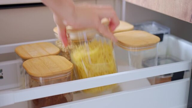 Hand opens pull out kitchen drawer, removes clear container filled with dry pasta and slides drawer closed. Concept of organized food storage and easy pantry access