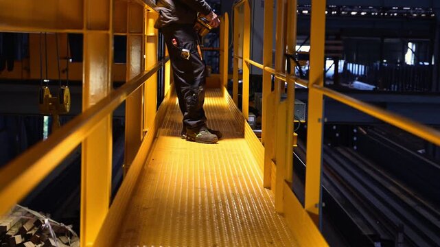 Industrial worker operating an overhead crane with a remote control from an elevated platform inside a heavy industrial facility, with his face completely unrecognizable.
