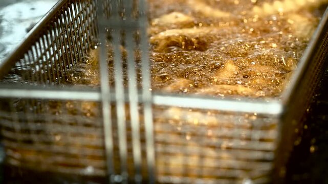 Close up of bubbling oil in industrial deep fryer with metal basket submerged, golden brown fries sizzling under warm amber kitchen light, steam rising in busy restaurant kitchen