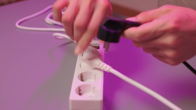 Neat electrical connection demonstration. Hands carefully connecting cords to power source. Detailed scene of person organizing wires on colorful illuminated desk surface