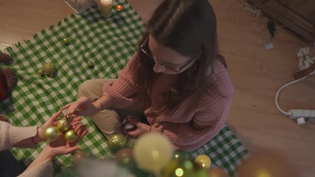 During a cozy Christmas activity, careful hands choose and attach golden ornaments to the lower branches of a tree. Overhead close view presents hands-on tree decorating with strong commercial appeal.