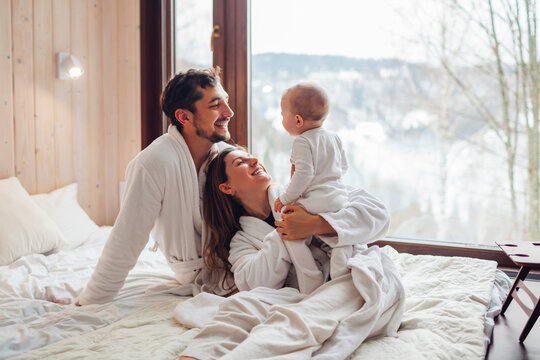 Happy family playing on bed enjoying snowy mountains landscape from hotel window. Mom and dad having fun with toddler baby girl