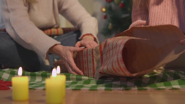 Friends wrap a present together on the floor beside warm candles during the holiday season. Close lifestyle view presents cooperative gift making with strong commercial appeal.
