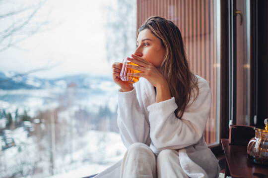 Drinking hot tea enjoying winter landscape in mountains. Woman wears bathrobe relaxing by big window of cabin house on holiday