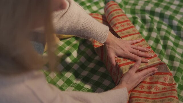 Close footage shows female hands that fold patterned paper neatly around a rectangular holiday gift. Overhead close view highlights careful present wrapping through warm seasonal detail.