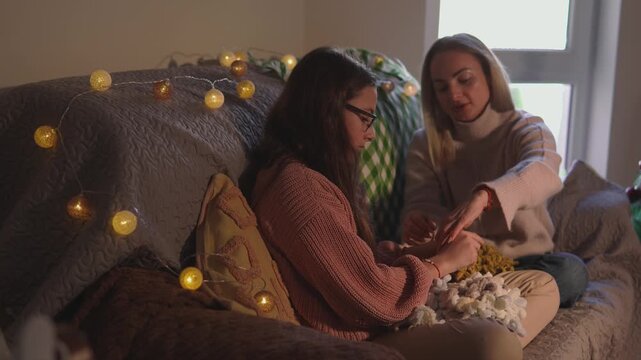 Two young women smile and share a playful moment with handmade holiday decorations in a festive home. Intimate portrait view captures genuine friendship for authentic Christmas storytelling.