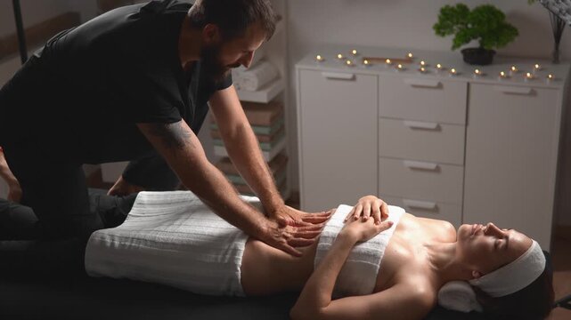 A client relaxes as a massage therapist provides a full abdominal massage while a woman rests beneath a towel. This treatment shows spa relaxation, body confidence and wellness practice.
