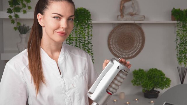 In a wellness session, a massage therapist presents a handheld device for professional spa body care. The therapist portrait view highlights aesthetic technology, wellness service and client care.