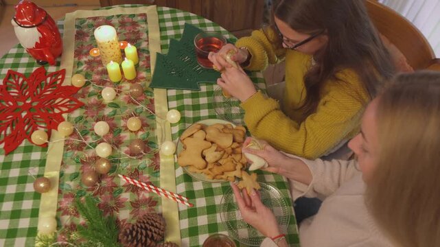 Two female friends arrange cookies, candles and ornaments across a richly decorated holiday table. Overhead lifestyle view highlights creative holiday preparation through warm seasonal detail.