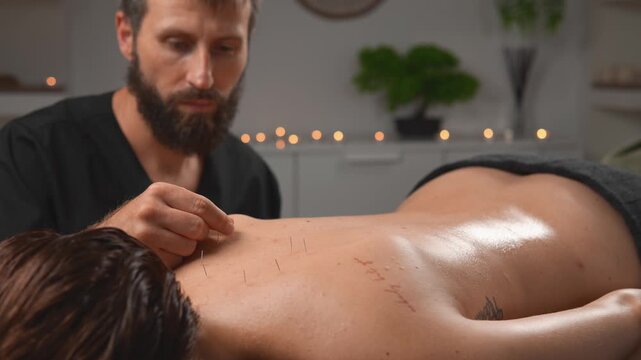 In a wellness session, a massage therapist monitors acupuncture needles during a calm studio session. The wide practitioner view highlights mind body balance, relaxation and client care.