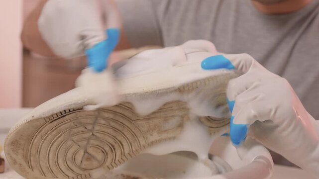 Homeowner cleans sneaker soles carefully. Sponge removes dirt from textured shoe soles. Closeup of homeowner washing sneaker soles using sponge and soapy water in sink