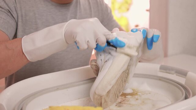 Homeowner cleans sneaker soles carefully. Sponge removes dirt from textured shoe soles. Closeup of homeowner washing sneaker soles using sponge and soapy water in sink