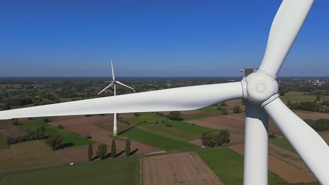 Static drone footage showing a close-up view of a wind turbine hub and blades in the foreground, with a second turbine visible farther away in the Belgian countryside