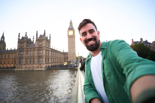 Smiling Caucasian millennial man taking a selfie beside the River Thames in London, with Big Ben and the Houses of Parliament behind. Wide-angle travel portrait conveying joy and sightseeing.