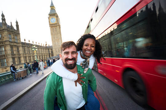 Joyful interracial couple having fun in London, with Big Ben and a red double decker bus behind. Playful piggyback pose, wide angle travel portrait that radiates energy and spontaneity.
