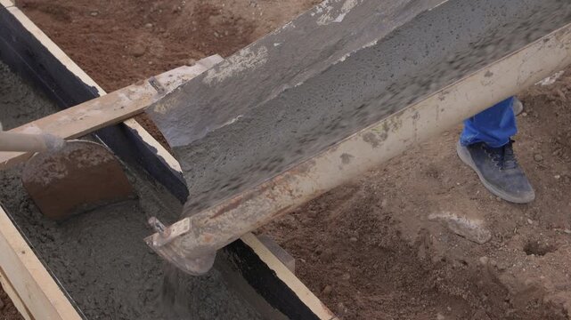 Construction worker actively pouring concrete into forms. Construction worker skillfully pouring wet concrete into formwork amid surrounding earth and safety gear