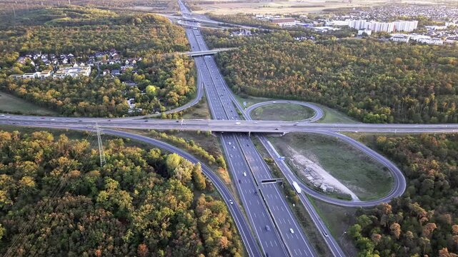 High oblique aerial view of multi level highway interchange with overpasses, ramps, and busy traffic through mixed woodland and suburban edge with looping cloverleaf connectors