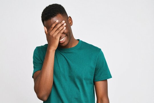 Young black man wearing green t-shirt makes facepalm gesture on white background. Handsome african american male laughs, smiles covering eyes with hand. Person feels embarrassed amused by silly