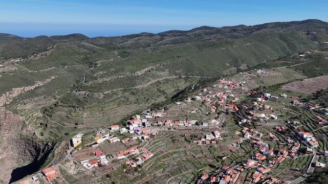 Aerial pull back drone shot revealing the beautiful remote El Cercado hamlet nestled in lush green hills near Punta del Arenero mountain on La Gomera (Canary Islands), with distant ocean views.