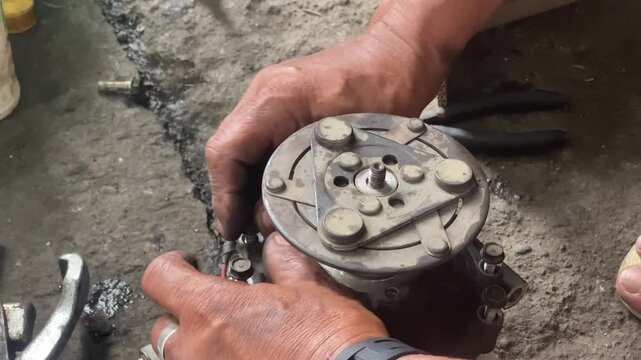 technician is assembling pieces of a broken car air conditioning compressor on the pavement.