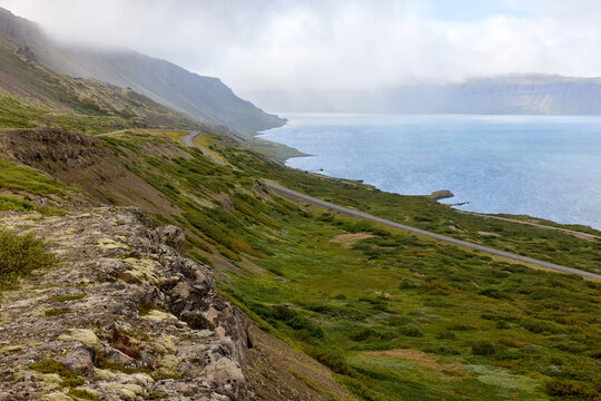 Road winding along misty fjord coast below steep slopes, Iceland