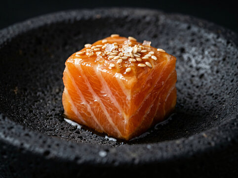 A single cube of fresh raw salmon sashimi topped with sesame seeds on a black stone plate.