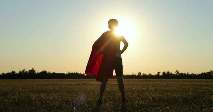 Boy superhero silhouette stands in field at sunset. Child wears red cape and imagines heroic adventure. Golden sunlight creates inspiring freedom and courage mood for cinematic storytelling.