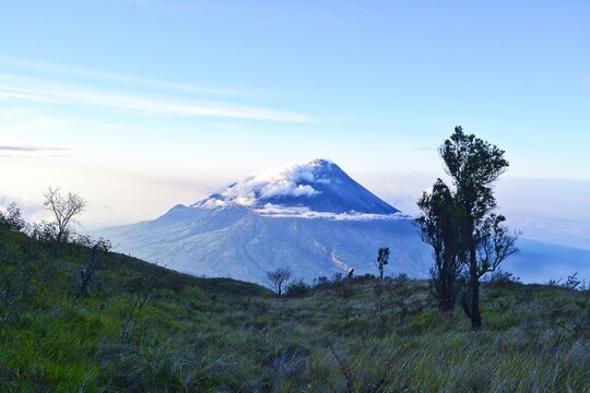 Majestic volcano peak with clouds at summit viewed from green grassy hill, clear blue sky morning landscape in Indonesia.