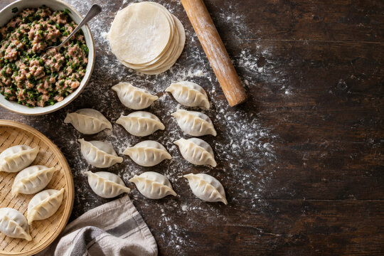 Overhead view of Lunar New Year dumpling making in progress with pleated raw dumplings, round wrappers, pork and chive filling and a rolling pin on a floured dark wooden table