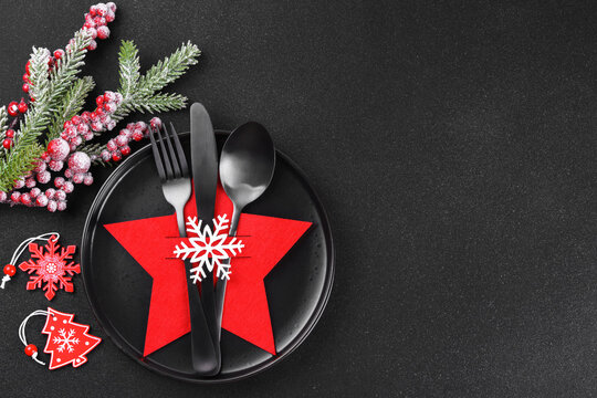 Holiday table setting with black plate and black cutlery in red star shaped cutlery holder with white snowflake and frosted Christmas tree branch on dark textured background, copy space.