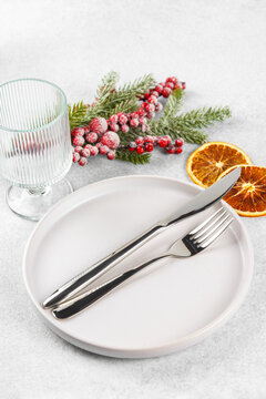 Festive holiday table setting with silver cutlery arranged on white plate with showy Christmas tree branch with red holly berry, dry orange and tall glass on light gray textured background