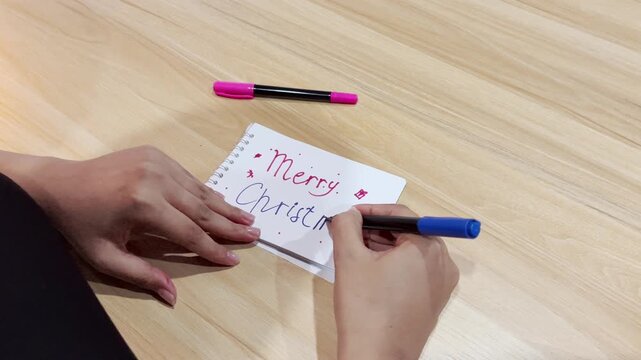Close-up of a women writing "Merry Christmas" greeting on a small white spiral notebook using a blue marker pen on a light wooden desk, Perfect for holiday, Christmas, and celebration