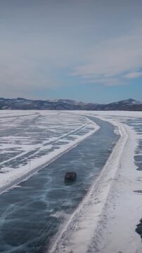 Drone footage of a vehicle driving along a cleared ice road on frozen Lake Baikal in winter, with snowy shores, mountains on horizon, and blue sky above