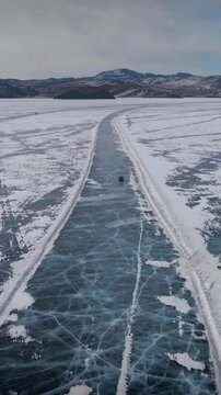 Drone view of a winter ice road across frozen Lake Baikal with blue cracks and snow patterns, with distant mountains under a cloudy sky, Siberia, Russia