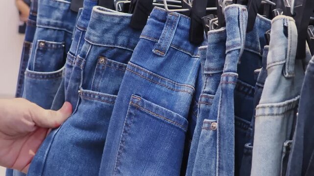 Man browsing blue jeans on clothing rack in store. Close-up of a customer checking denim pants by their fabric. Concept of menswear shopping, casual fashion and consumer choice.