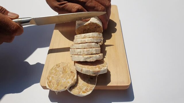 Close up of hands slicing raw tempeh or tempe on a wooden cutting board against a white background.