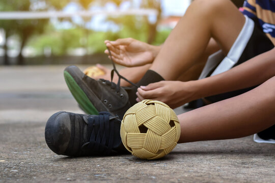 A sepak takraw player sits on the ground, tying their shoelaces next to a woven rattan ball.
