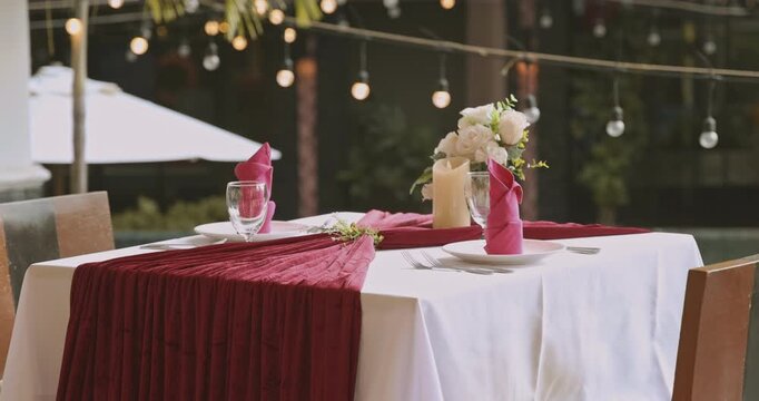 Romantic dinner table setting with red napkins and flowers. Elegant restaurant dining table for two with warm string lights in background. Cozy evening atmosphere for anniversary or date night.