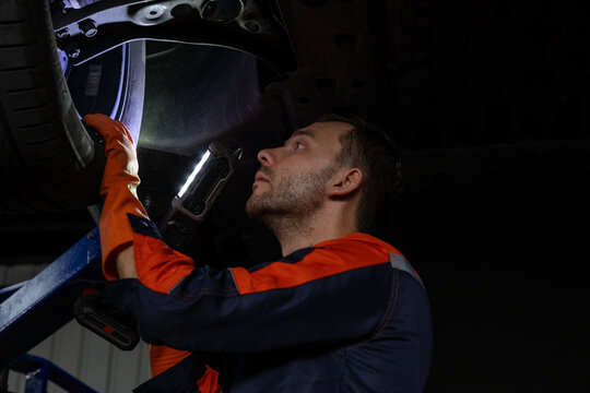 Mechanic in orange protective clothing inspects vehicle suspension inside dark garage using handheld work light with focused attention and safety care