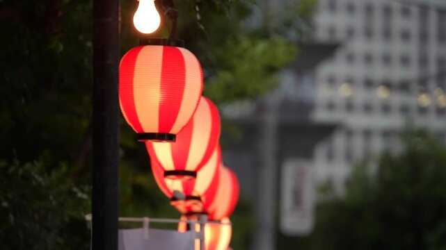 cinematic shallow depth of field shot of illuminated traditional japanese red and white paper lanterns hanging in a row along a city street at dusk
