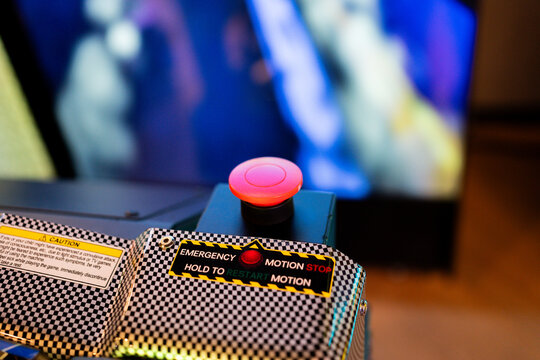 Close-up shot of a red emergency motion stop button on an arcade simulator machine, showing clear safety warning stickers, caution labels, and restart instructions in an amusement arcade venue.