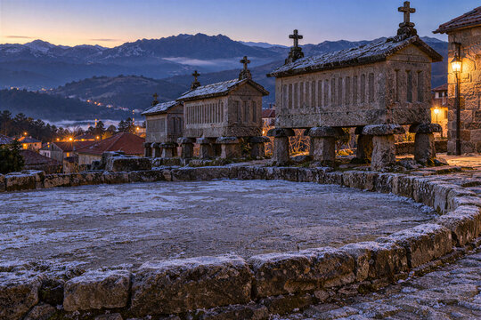 Row of stone granaries around a communal threshing floor at blue hour with village lights
