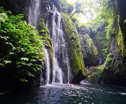 Tall mossy waterfall cascading into natural pool in lush green jungle canyon, person floating relaxed, tropical paradise in Indonesia.