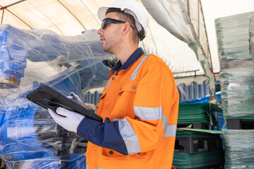 Industrial worker wearing orange safety uniform and helmet, inspecting wrapped cargo with digital tablet inside warehouse storage area © 220 Selfmade studio
