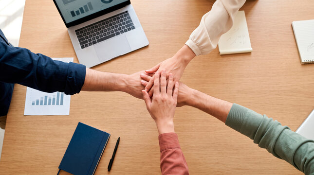 Business colleagues joining hands over a wooden desk with a laptop and financial charts to symbolize unity and corporate teamwork