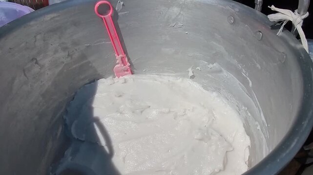 Video of Hand stirs Indonesian sweet coconut rice porridge in large metal pot. Sunlight highlights the creamy texture of the batter. Surrounding containers hold colorful liquids and tools. 4K Cuisine